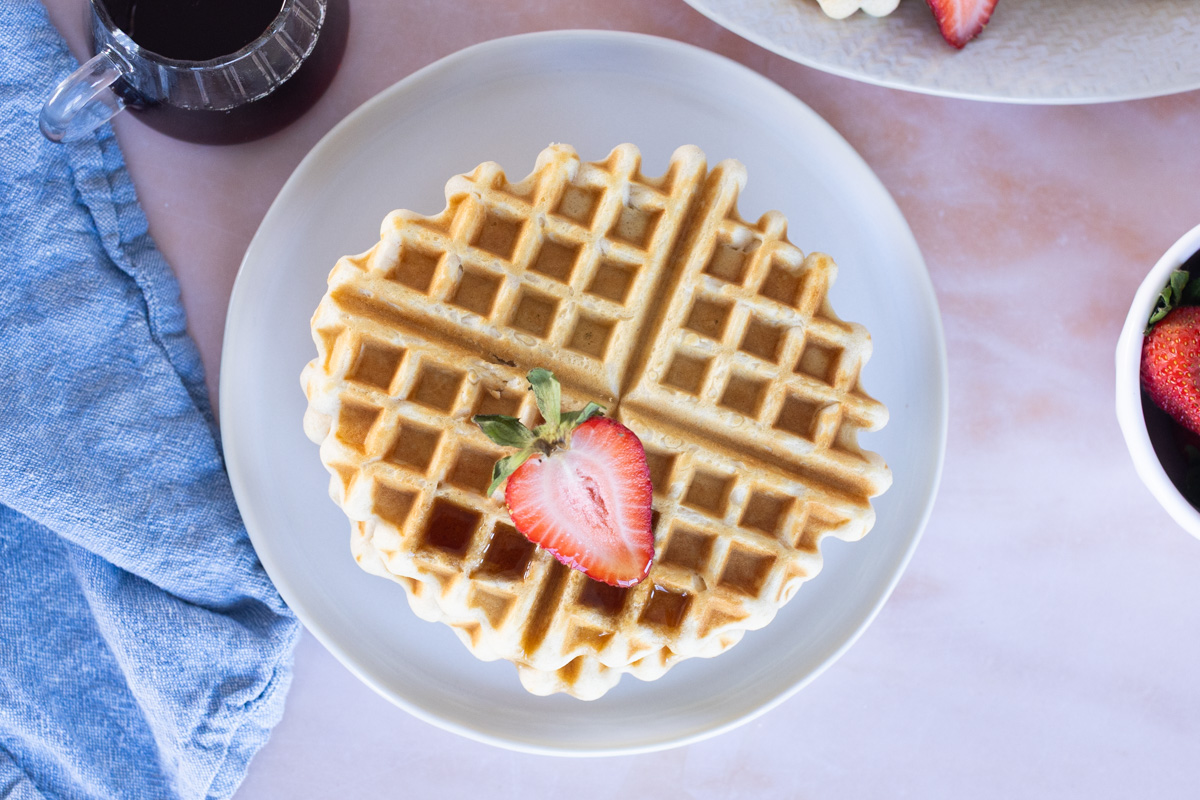 A round Easy Waffle on a white plate topped with sliced strawberry, served with syrup, a blue napkin, and a bowl of strawberries nearby.