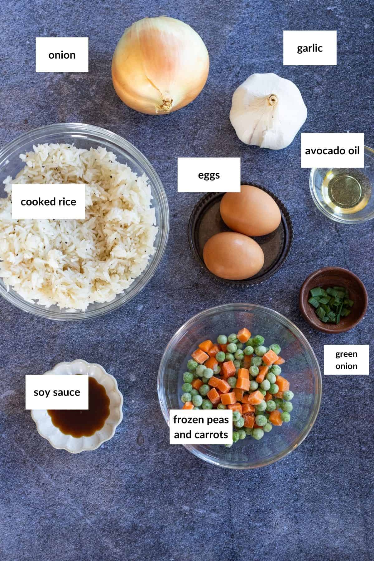 Top-down view of ingredients in small glass bowls: onion, garlic, avocado oil, 2 whole eggs, chopped green onion, cooked rice, soy sauce, and a bowl of frozen peas and carrots.