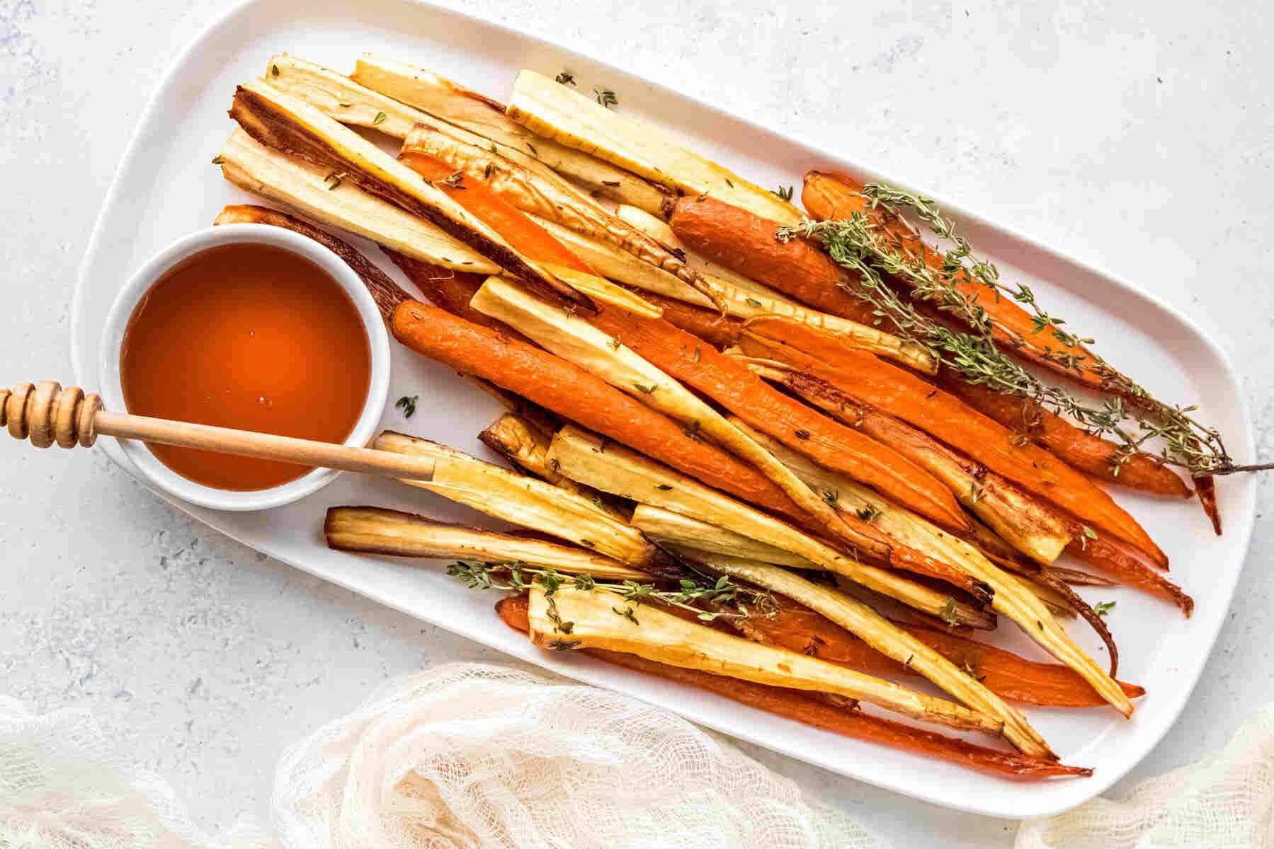 A white platter with roasted carrots and parsnips garnished with fresh thyme, served with a small bowl of honey and a honey dipper.