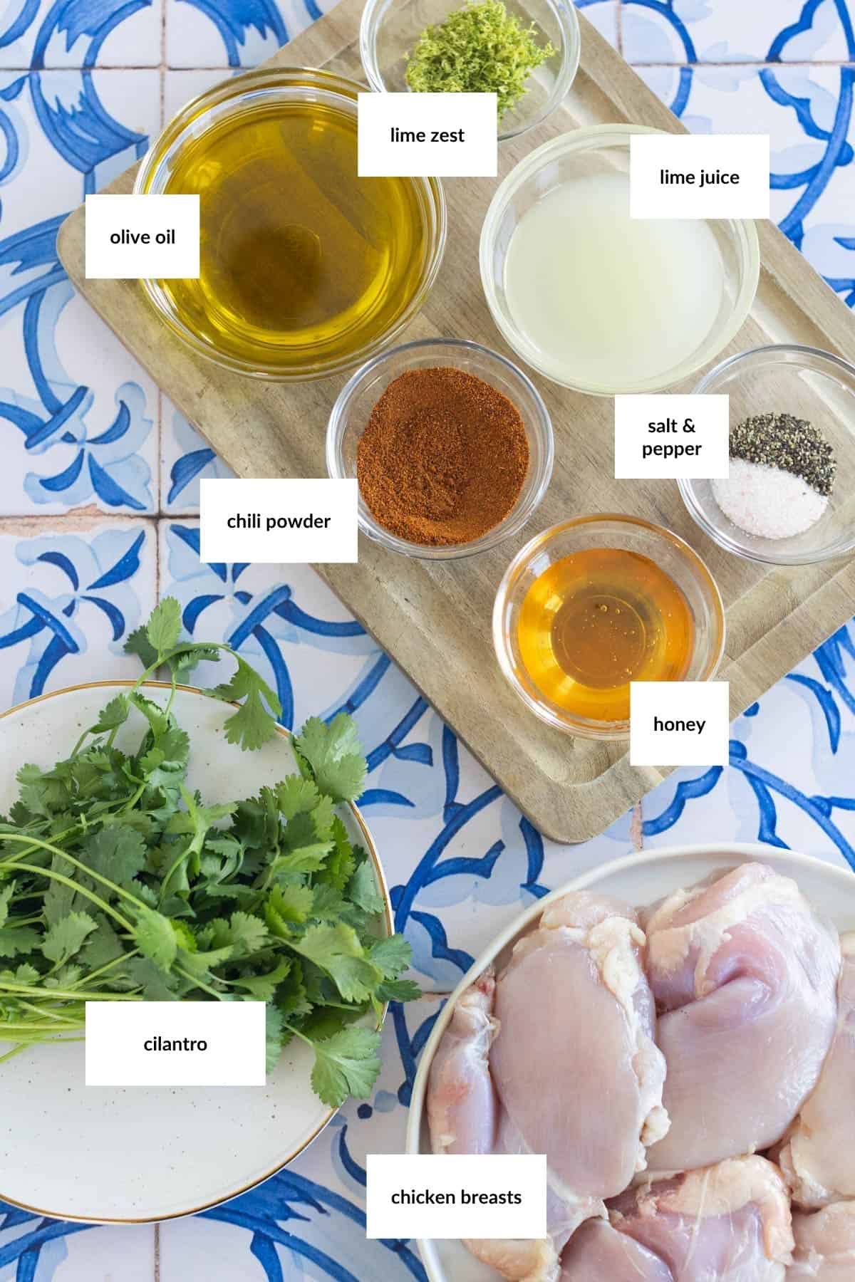 Ingredients for a recipe arranged on a patterned surface, including labeled bowls of olive oil, lime zest, lime juice, chili powder, salt & pepper, honey, a plate of cilantro, and raw chicken breasts.