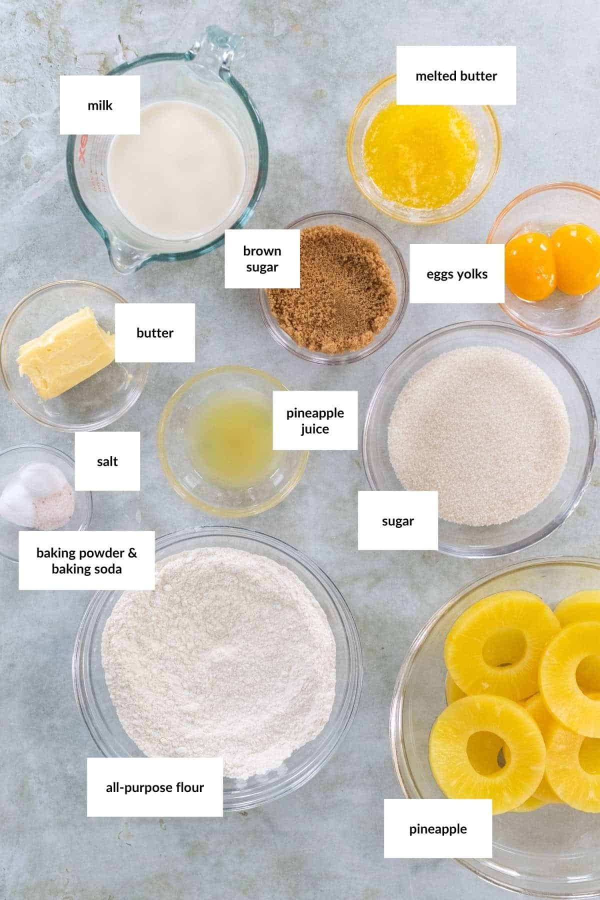 Overhead view of baking ingredients in bowls, including milk, melted butter, butter, brown sugar, egg yolks, salt, pineapple juice, sugar, flour, baking powder, and pineapple rings.