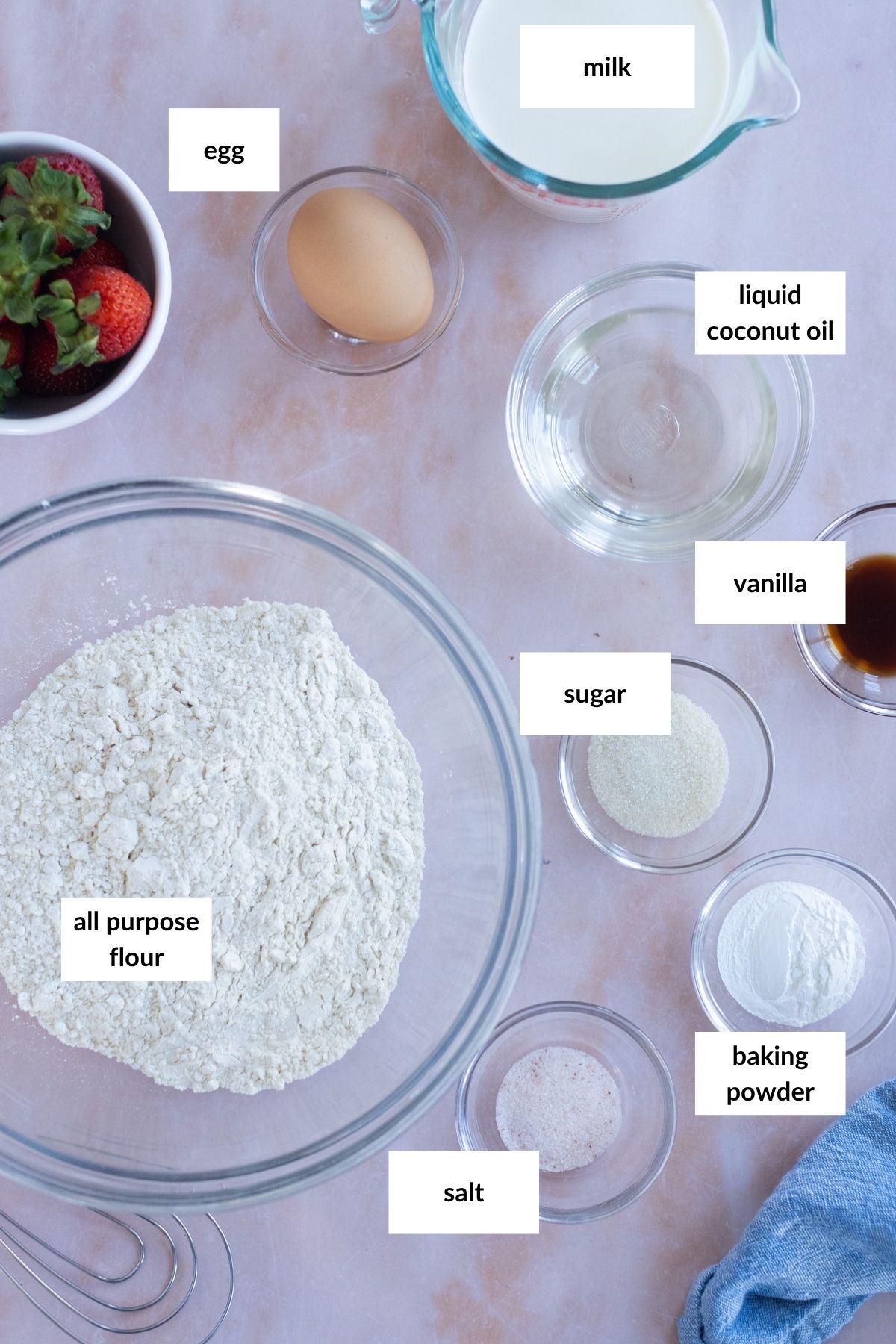 Baking ingredients on a countertop include all-purpose flour, egg, milk, liquid coconut oil, vanilla, sugar, baking powder, salt, and a bowl of strawberries.