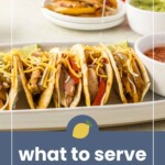 Four chicken fajita tacos topped with cheese are served on a platter, with salsa, guacamole, and sliced bell peppers in the background. A hand squeezes lime over the tacos.