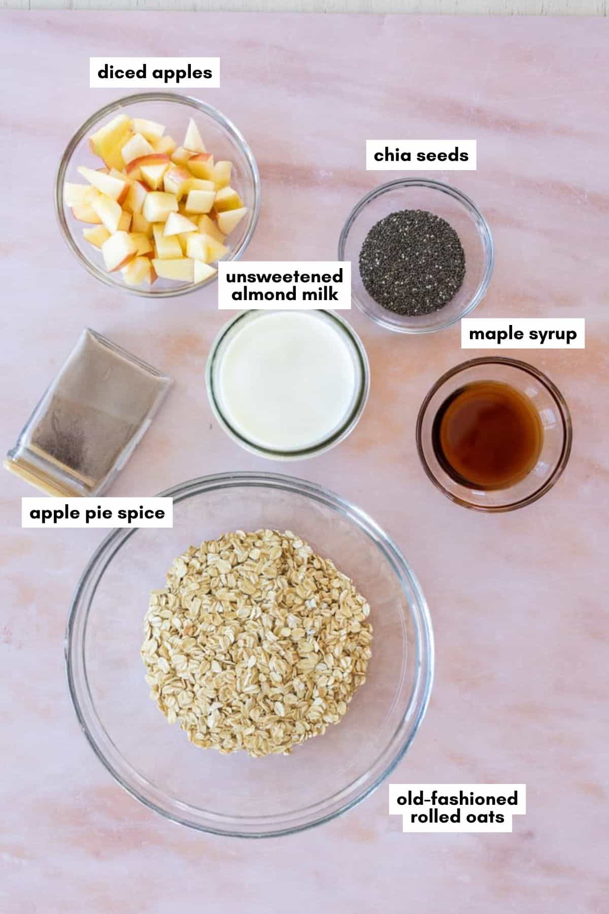 Top-down view of bowls containing diced apples, chia seeds, almond milk, maple syrup, apple pie spice, and rolled oats—each labeled with text—arranged on a light pink surface.