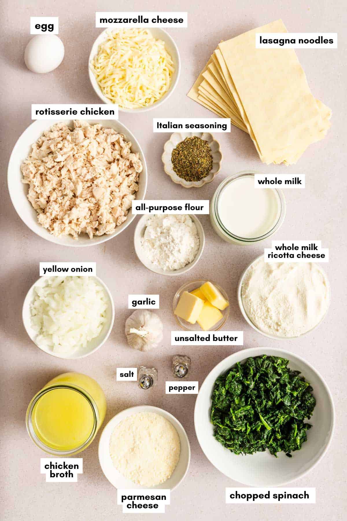 Various labeled ingredients in bowls and on a light colored surface including cheese, shredded chicken, lasagna noodles, spinach, broth, milk, butter, flour, spices, and an egg.