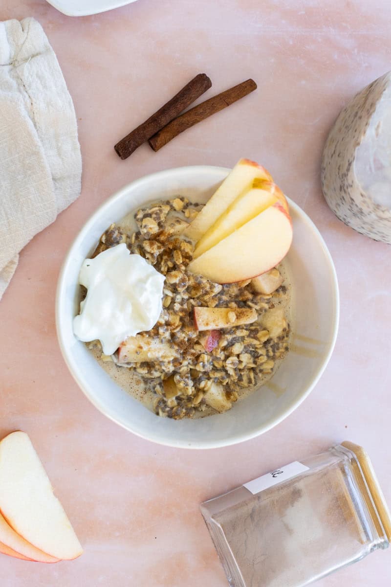 A bowl of oatmeal topped with apple slices and whipped cream sits on a pink surface, with cinnamon sticks and a jar of spices nearby.