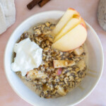 A bowl of oatmeal with apple slices, a dollop of yogurt, and a sprinkle of cinnamon, next to two cinnamon sticks on a pink surface.