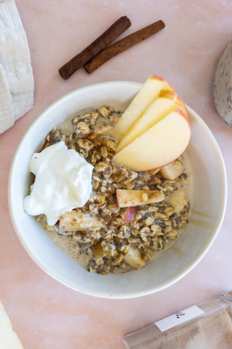 A bowl of oatmeal with apple slices, a dollop of yogurt, and a sprinkle of cinnamon, next to two cinnamon sticks on a pink surface.