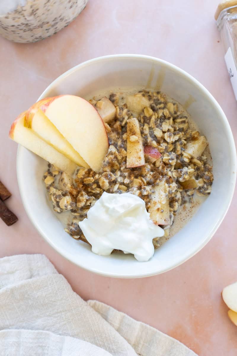 Bowl of apple cinnamon overnight oats sits on a pink surface topped with apple slices, a dollop of yogurt, and a sprinkle of cinnamon.