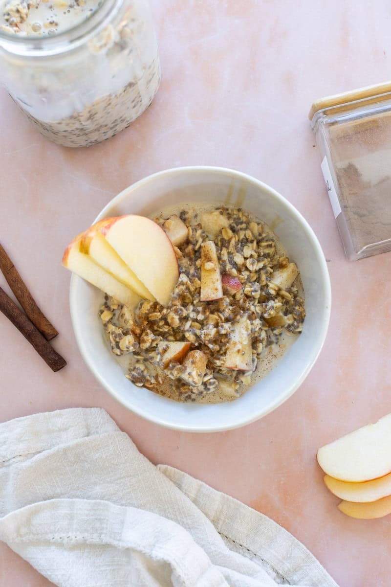 A bowl of overnight oats with apple slices, chia seeds, and cinnamon sits on a light pink surface next to a jar, cinnamon sticks, and a folded napkin.