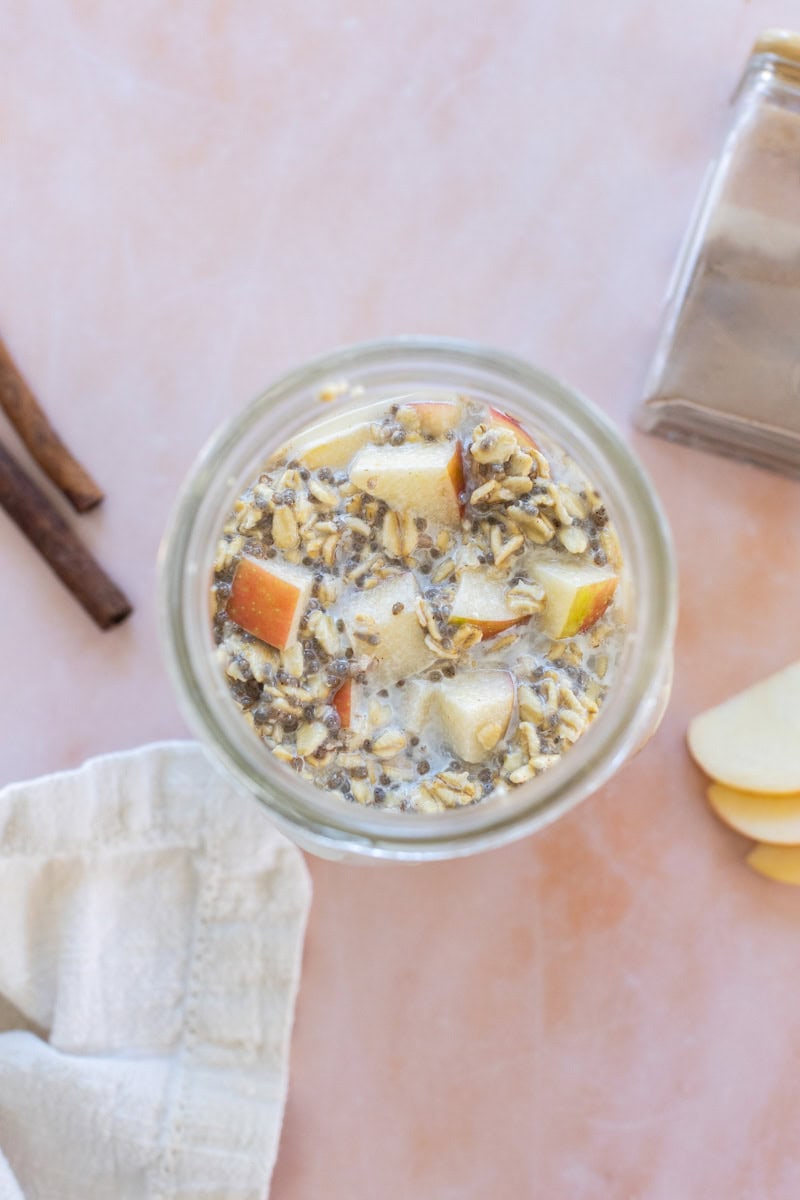 A glass jar filled with overnight oats, chia seeds, and apple pieces sits on a light surface beside cinnamon sticks, a spice jar, apple slices, and a white cloth.