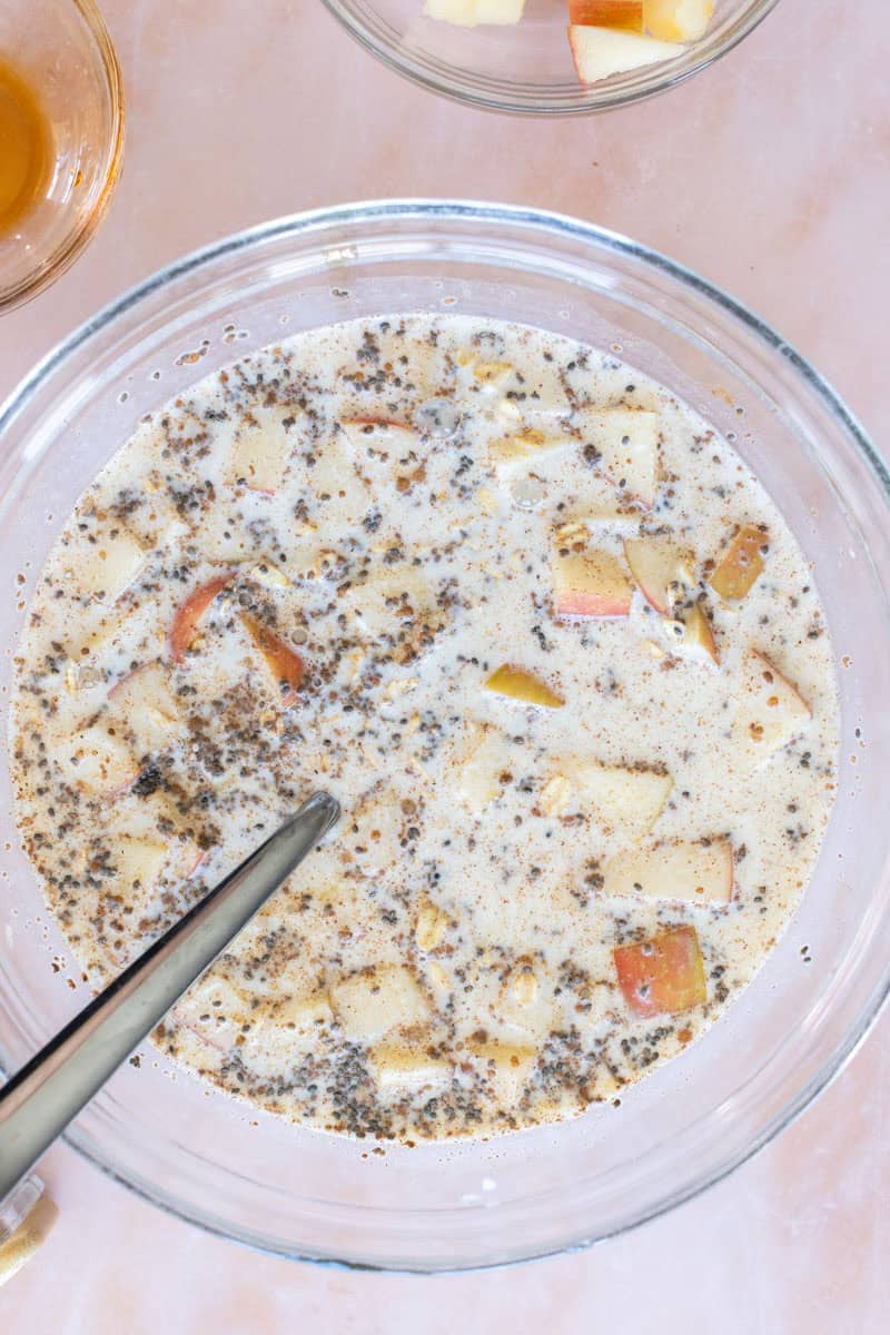 A glass bowl filled with a mixture of chopped apples, milk, chia seeds, and spices, with a spoon resting in the bowl.