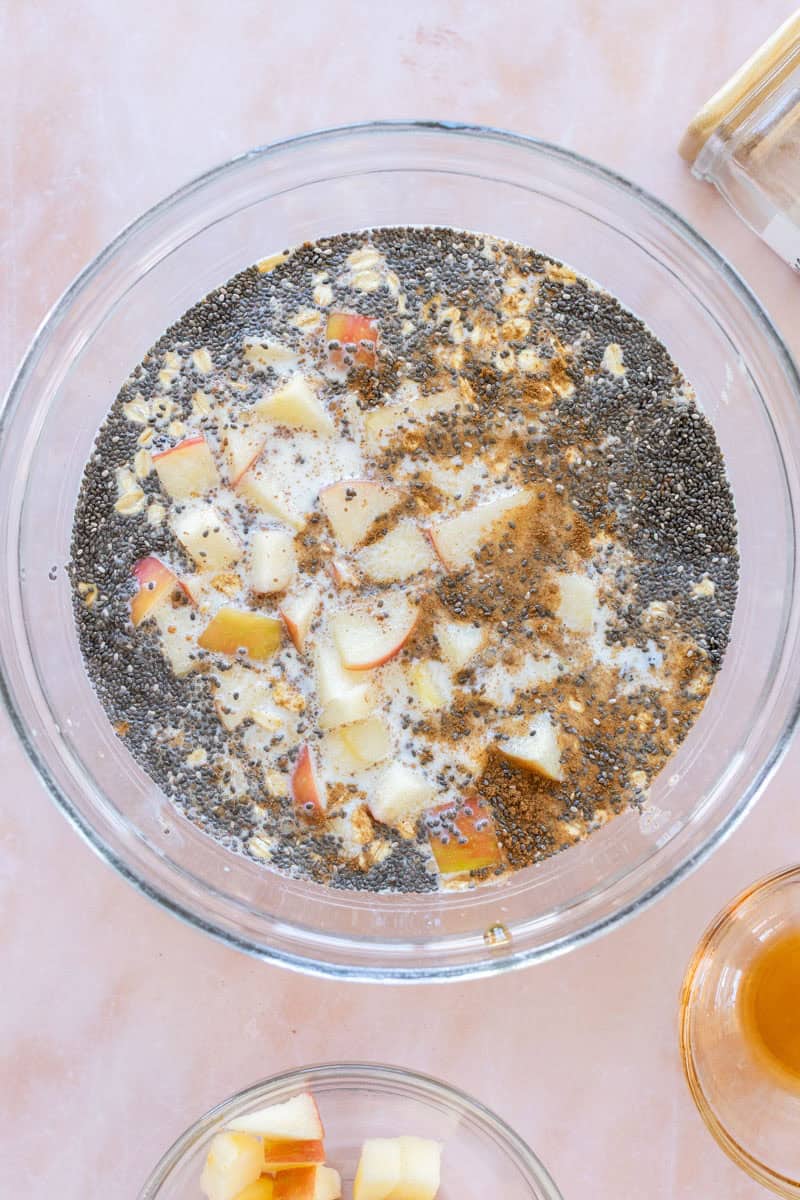 A glass bowl contains chia seeds, diced apples, oats, cinnamon, and milk on a light pink surface.