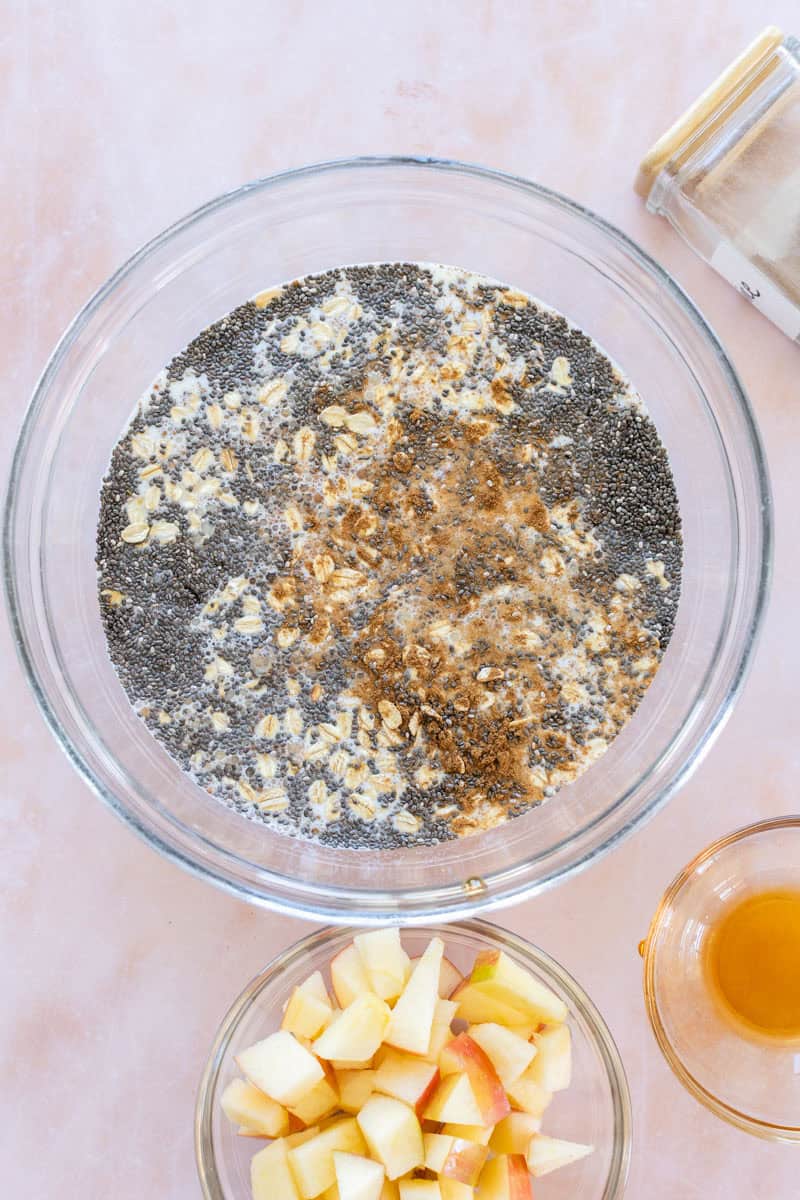 A glass bowl with oats, chia seeds, milk, and cinnamon, next to chopped apples and a small bowl of honey on a light surface.