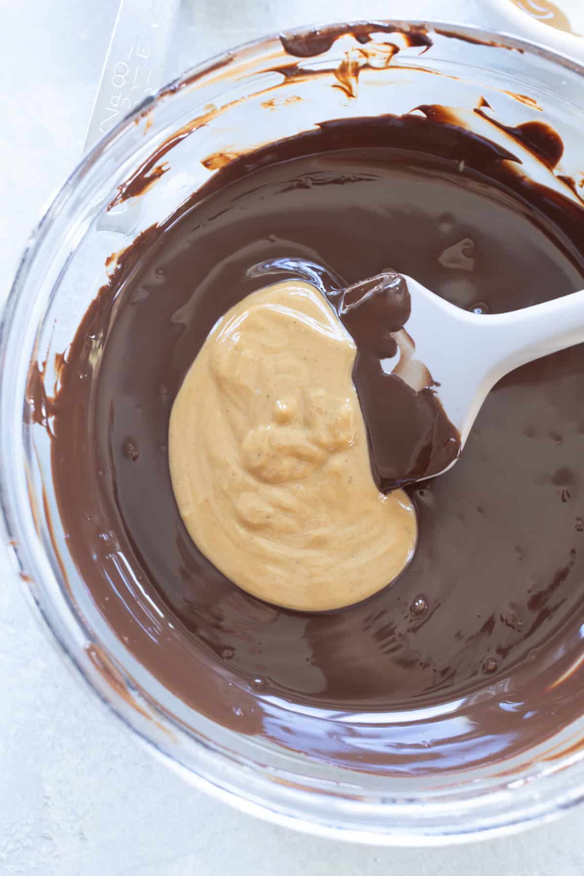 A glass bowl with melted chocolate and a dollop of peanut butter being mixed with a white spatula.