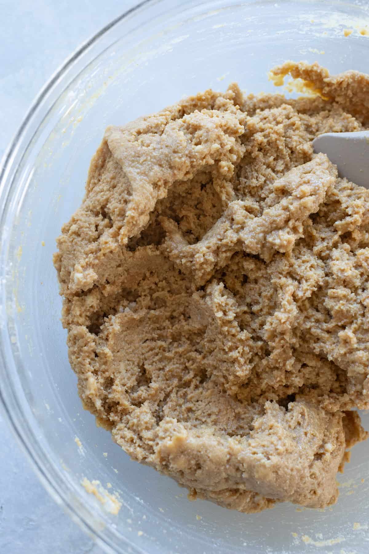 A clear glass bowl filled with light brown cookie dough being mixed with a spatula.