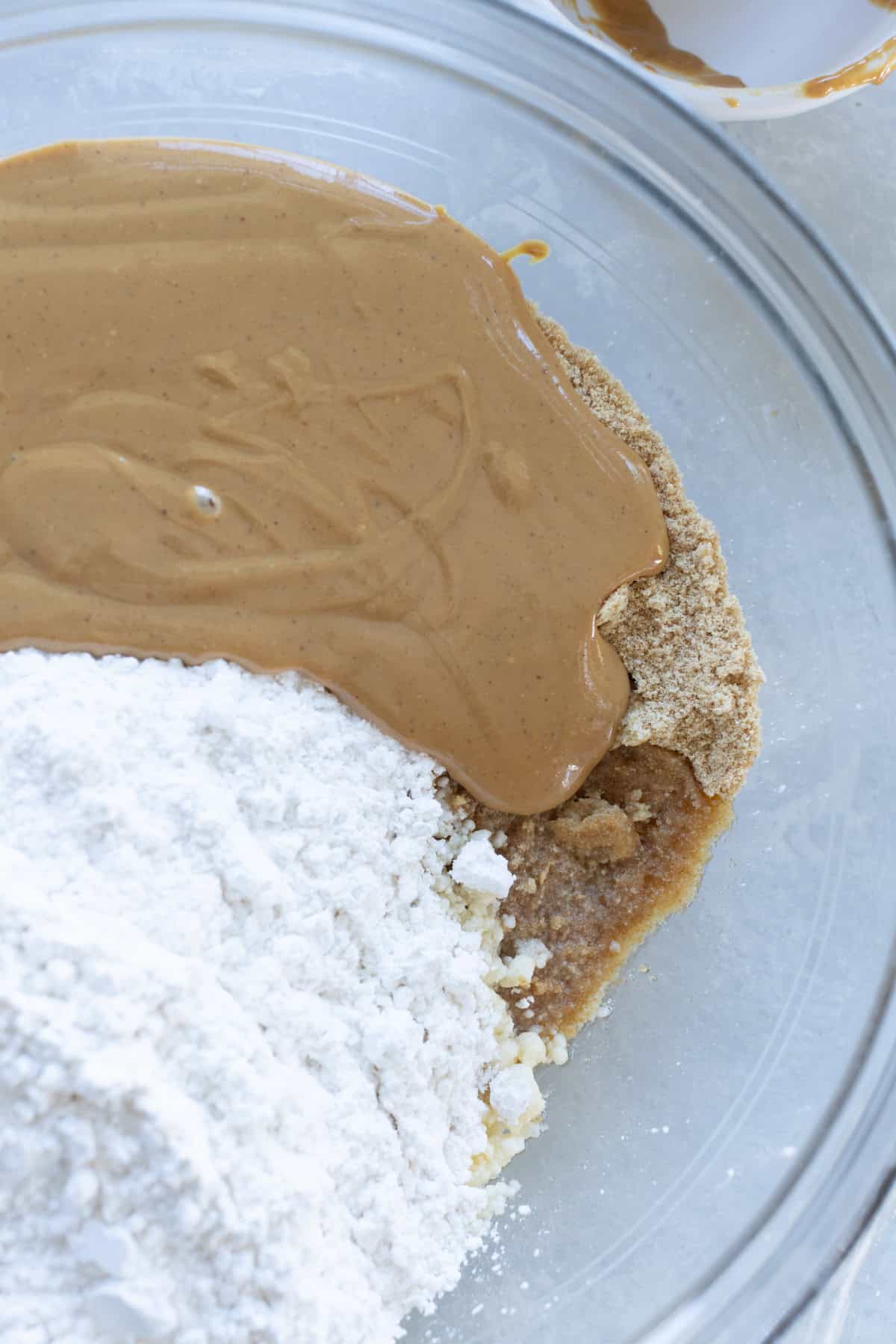 A glass bowl contains peanut butter, flour, and brown sugar, partially mixed together, viewed from above.