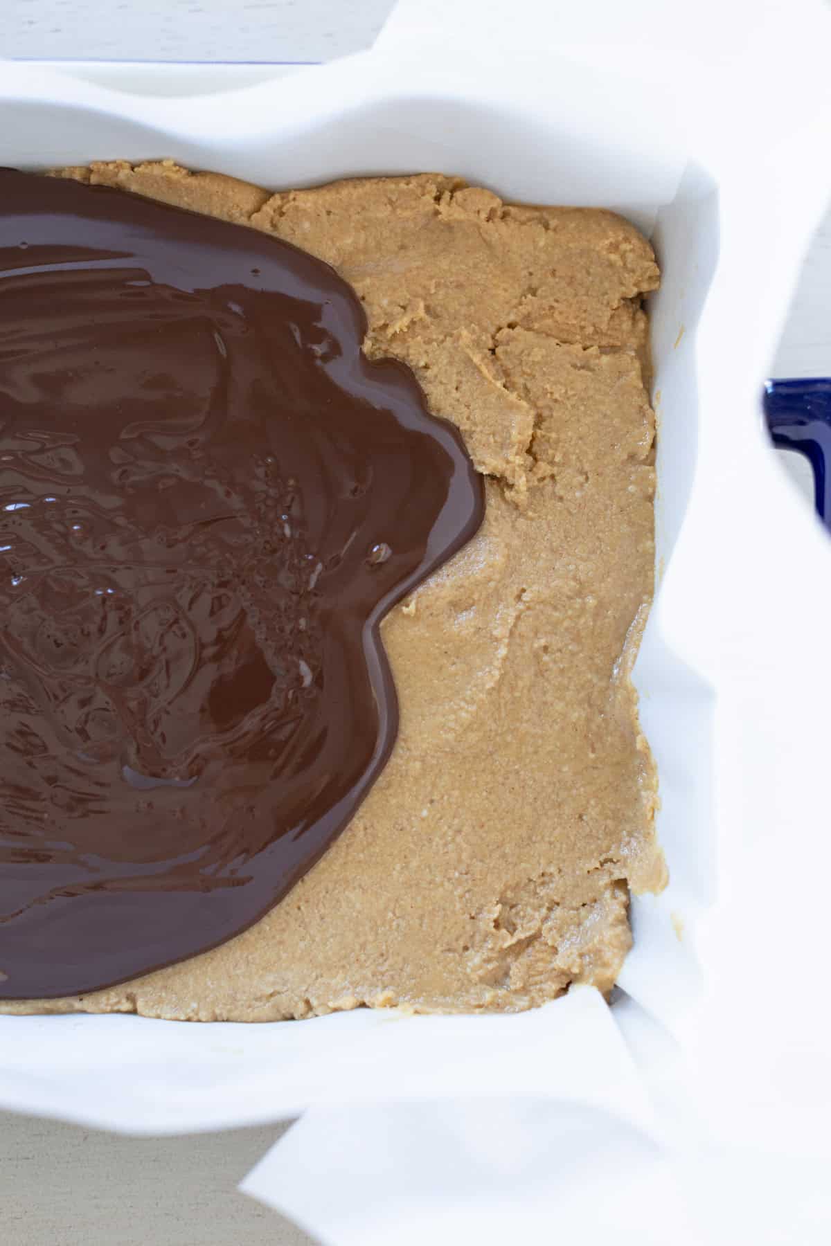 A baking dish lined with parchment paper holds a layer of peanut butter mixture, partially covered with melted chocolate.
