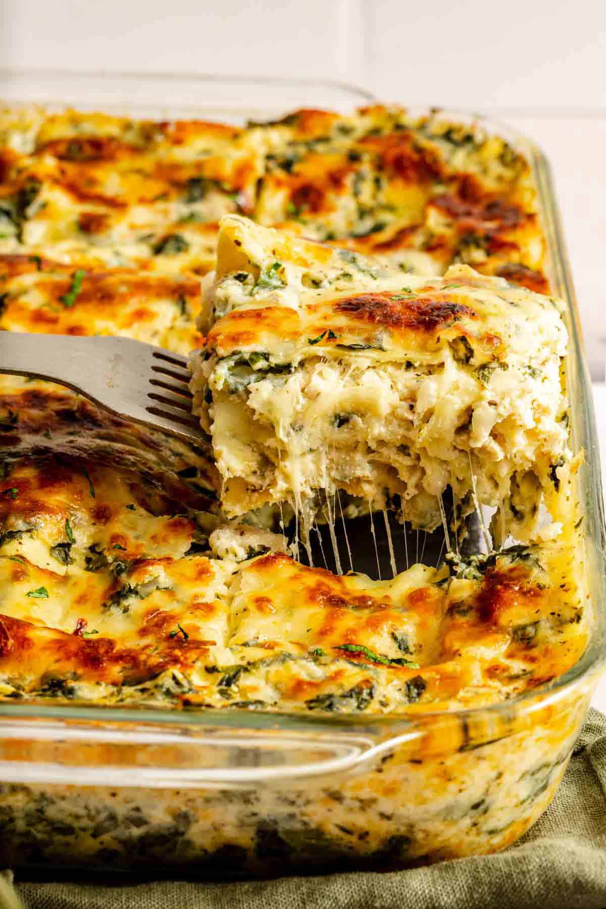 A metal spatula lifts a cheesy, golden-baked piece of spinach and cheese lasagna from a glass baking dish.