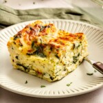 A slice of spinach and cheese lasagna on a beige plate with a fork, featuring layers of pasta, ricotta, spinach, and a golden-brown top.