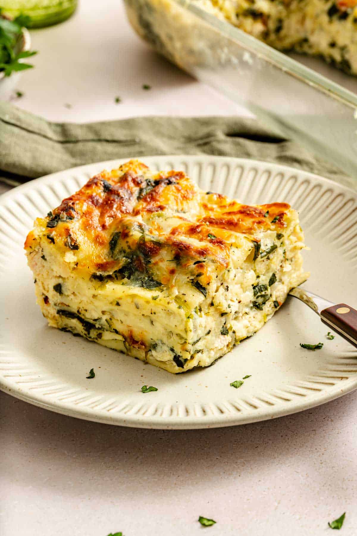 A slice of spinach and cheese lasagna on a beige plate with a fork, featuring layers of pasta, ricotta, spinach, and a golden-brown top.