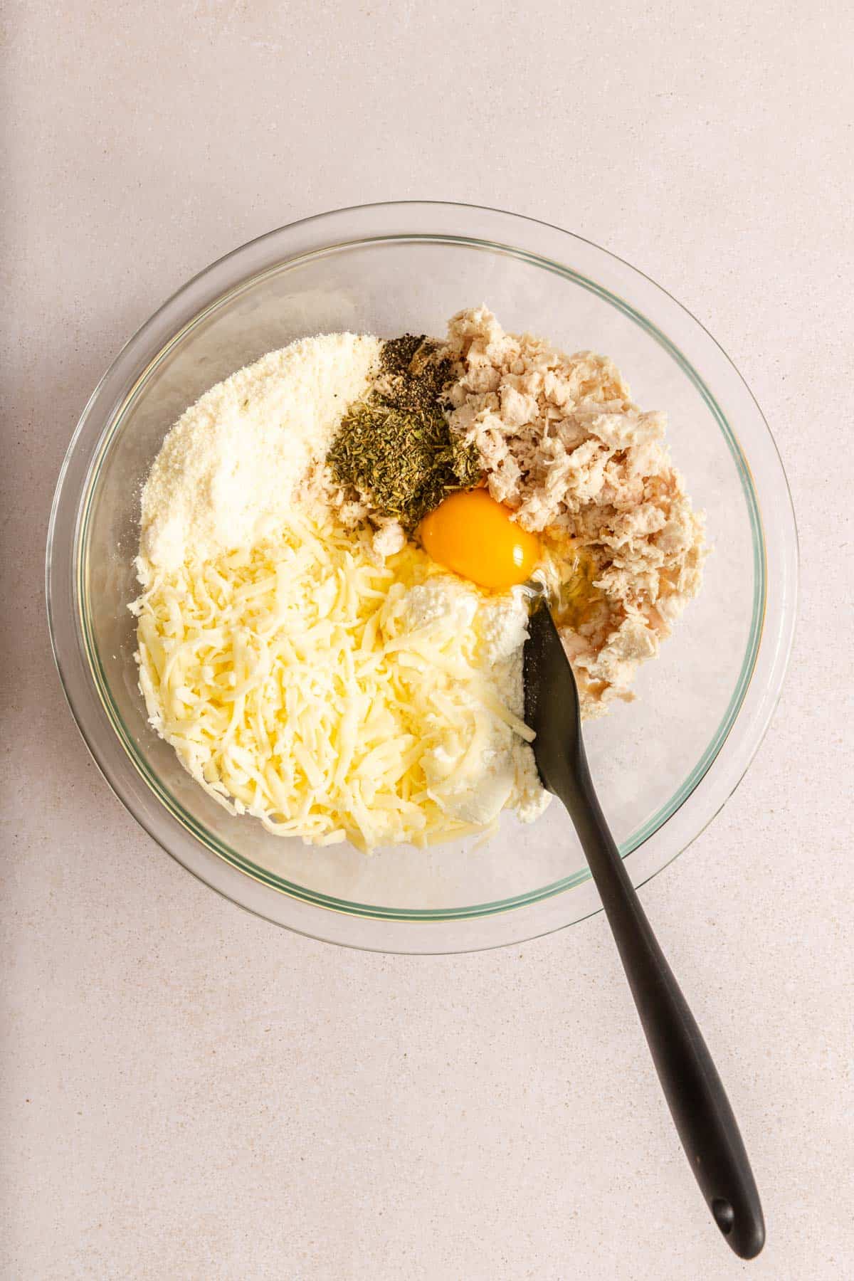 A clear glass bowl with shredded cheese, grated parmesan, an egg, herbs, tuna, and a spoon, all on a light countertop.