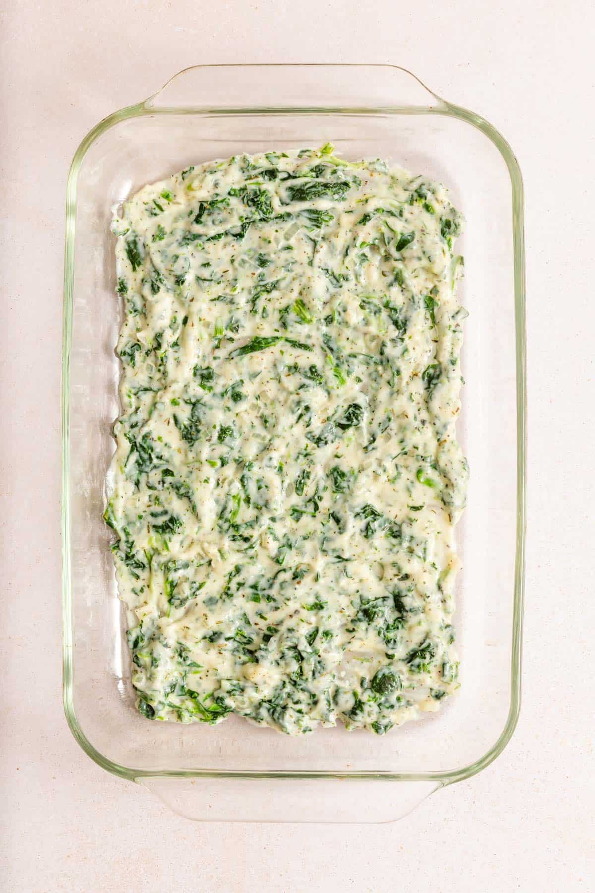 A glass baking dish filled with an uncooked mixture of spinach and cheese in a creamy white sauce, spread evenly.