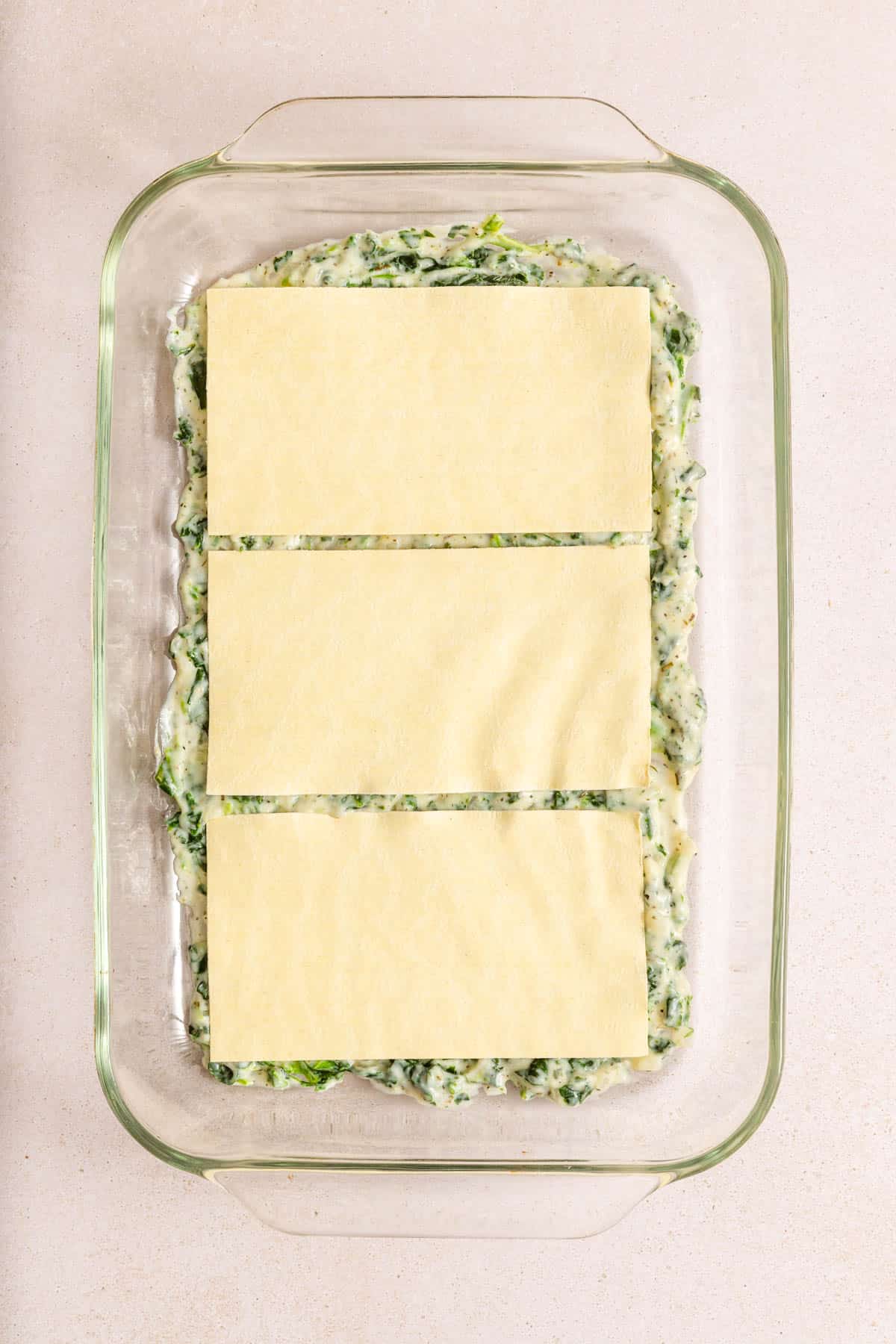 A glass baking dish containing a layer of spinach and cheese mixture topped with three uncooked lasagna noodles on a light countertop.