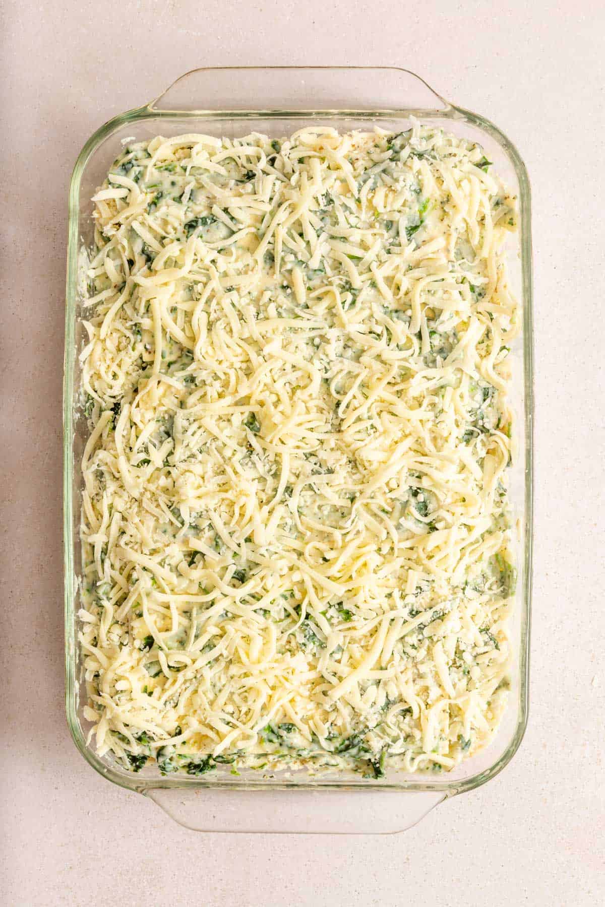 Rectangular glass baking dish filled with an uncooked creamy spinach casserole covered with a layer of shredded cheese on top.