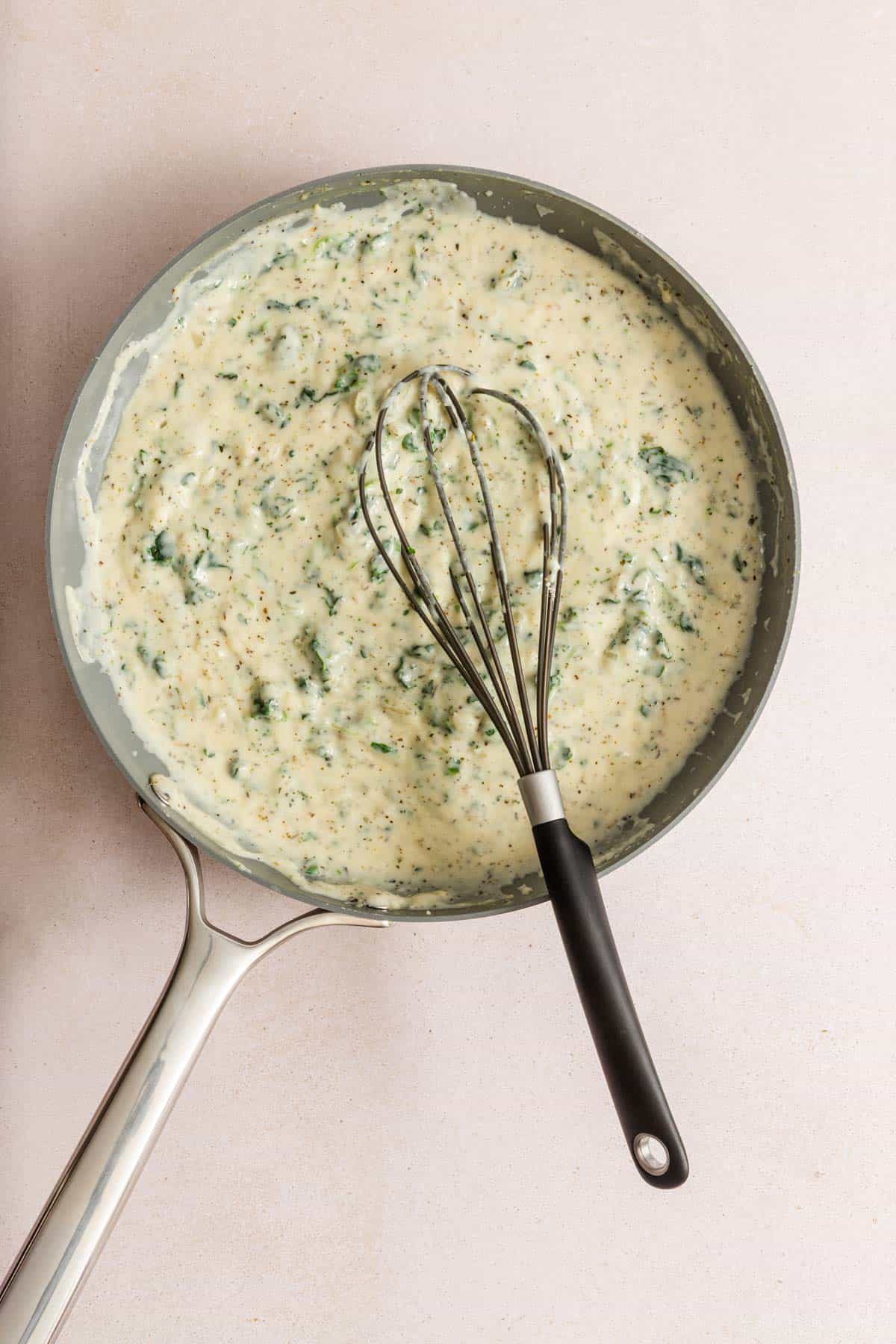 A saucepan filled with creamy spinach sauce, with a whisk resting inside, on a light-colored surface.