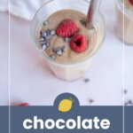 A glass of chocolate smoothie topped with two raspberries and chocolate chips, with a metal straw, on a light surface.