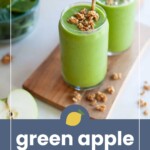 Two glasses of green smoothie with granola topping and metal straws on a wooden board, with spinach and apple slices nearby. Text reads “green apple smoothie.”.