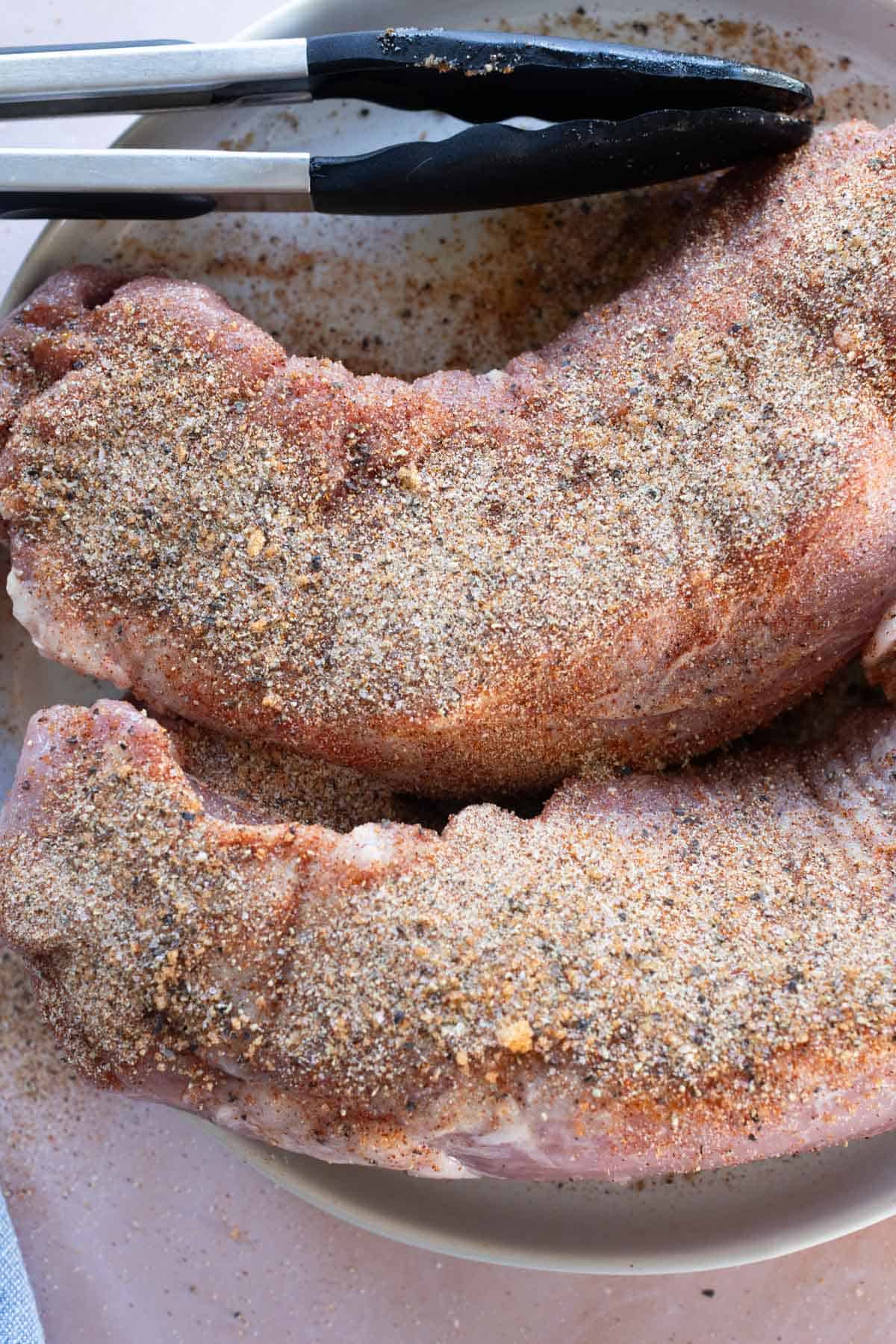 Two raw pork tenderloins coated with dry seasoning sit on a plate with metal tongs resting above them.