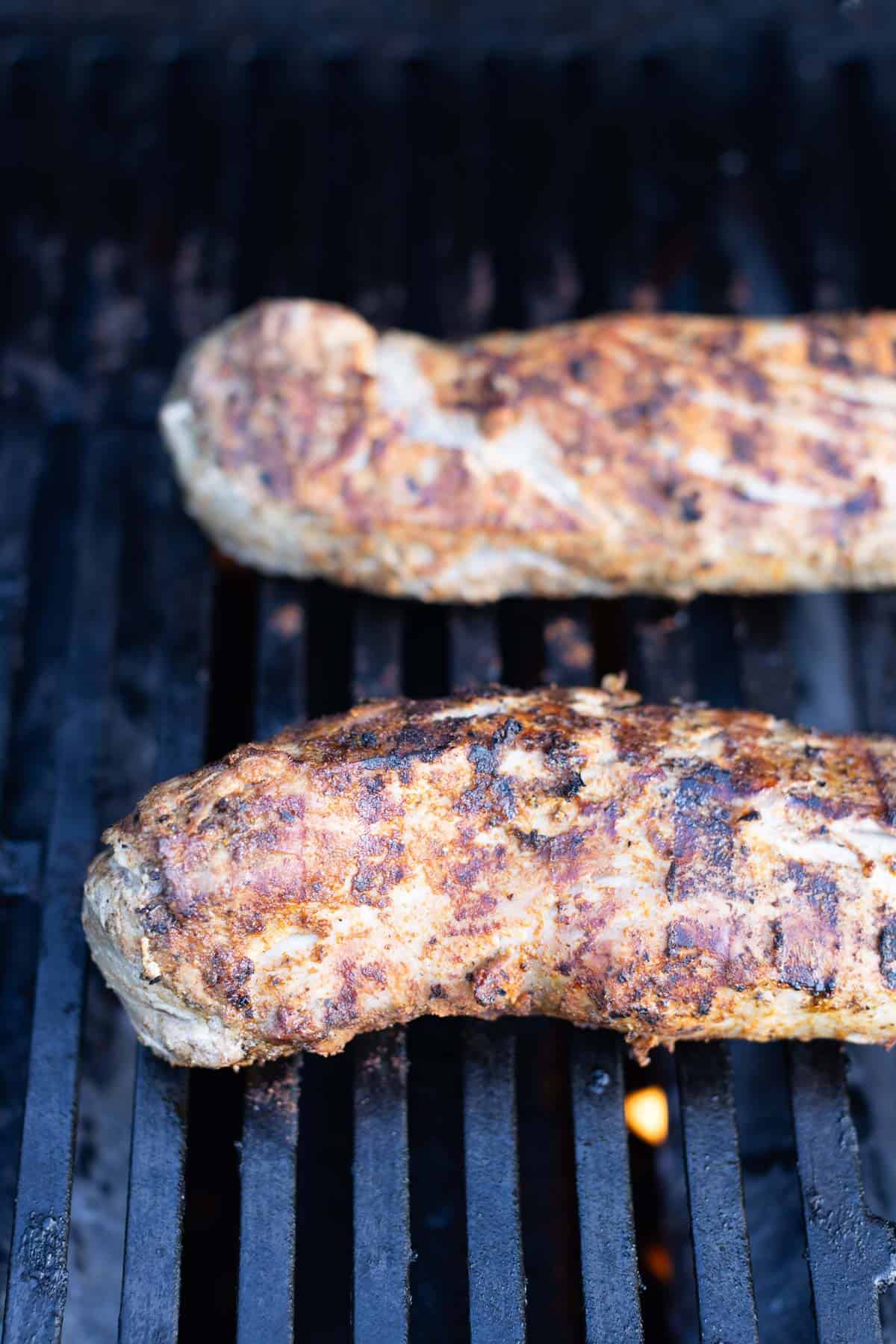Two seasoned pork tenderloins are cooking on a grill, showing grill marks and a lightly charred exterior.