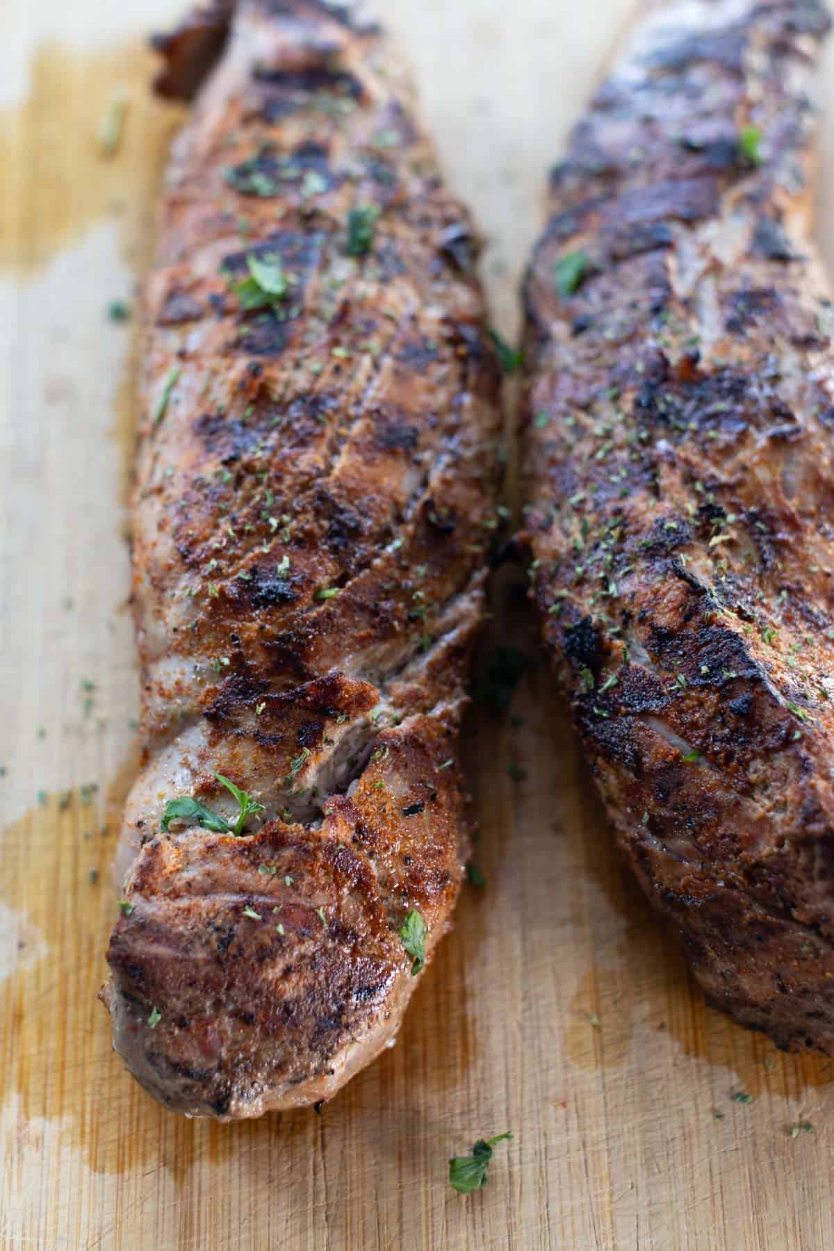 Two grilled pork tenderloins with grill marks and herbs, resting on a wooden cutting board.