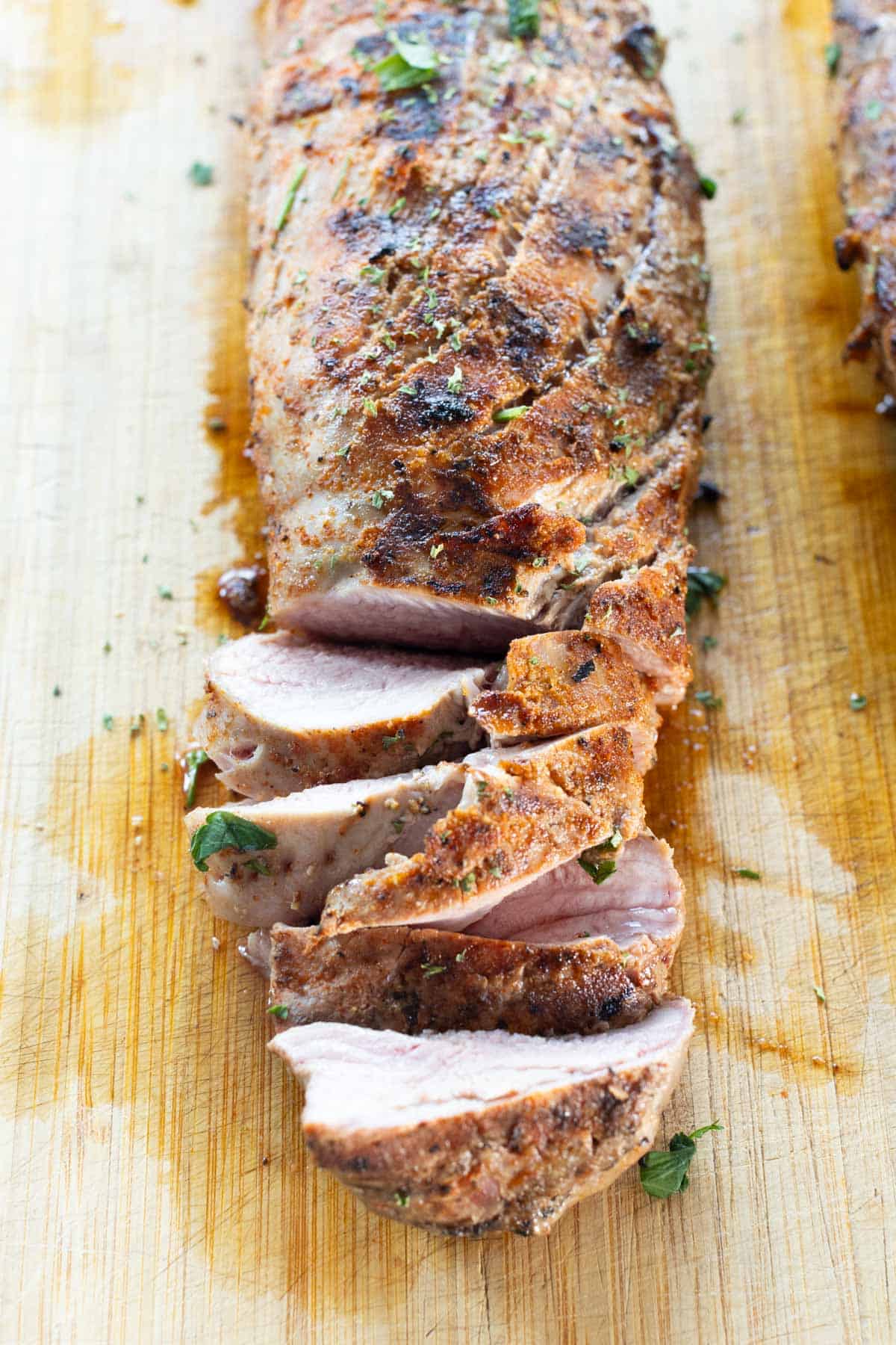 Sliced grilled pork tenderloin on a wooden cutting board, with visible seasoning and garnished with chopped herbs.