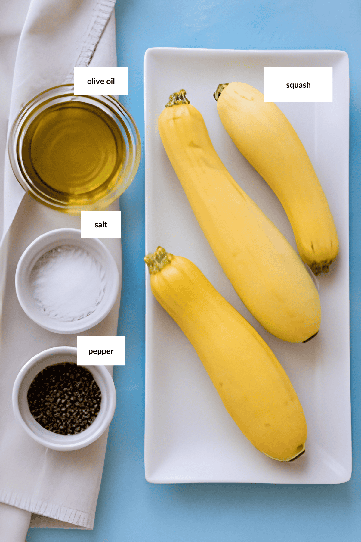 Three yellow squash on a rectangular plate, ready to become grilled squash, with bowls of olive oil, salt, and pepper, all labeled, arranged on a light blue surface with a napkin.