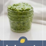 A small glass jar filled with green kale pesto sits on a white surface. The label below reads "kale pesto" with a lemon graphic above the text.