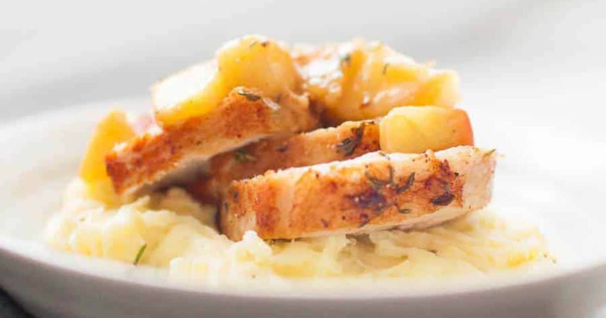 Sliced pork with apple topping served on mashed potatoes, presented on a white plate.