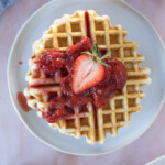 A plate with a waffle topped with strawberry sauce and a halved strawberry, shot from above on a light pink surface.