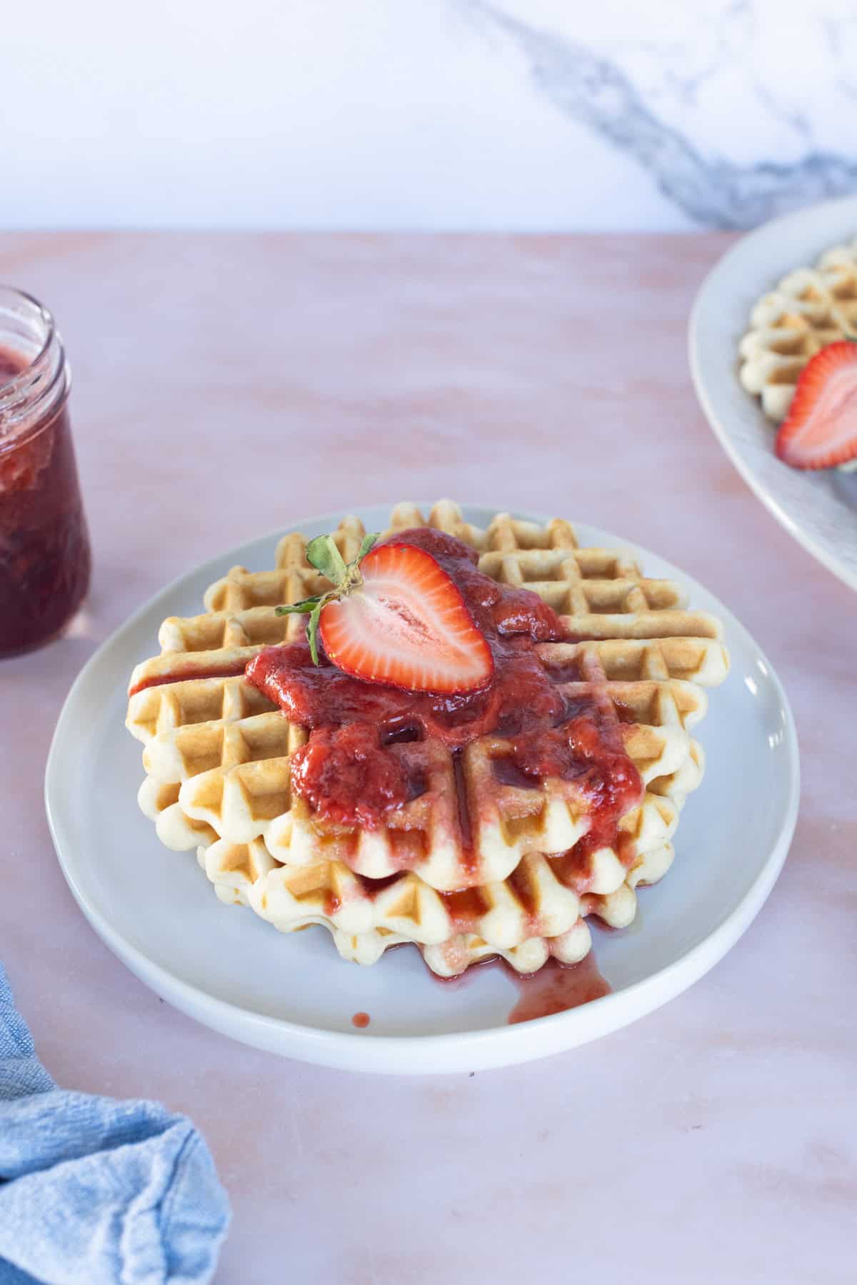 A plate with a waffle topped with strawberry sauce and a halved strawberry, next to a jar of sauce and another plate with waffles in the background.