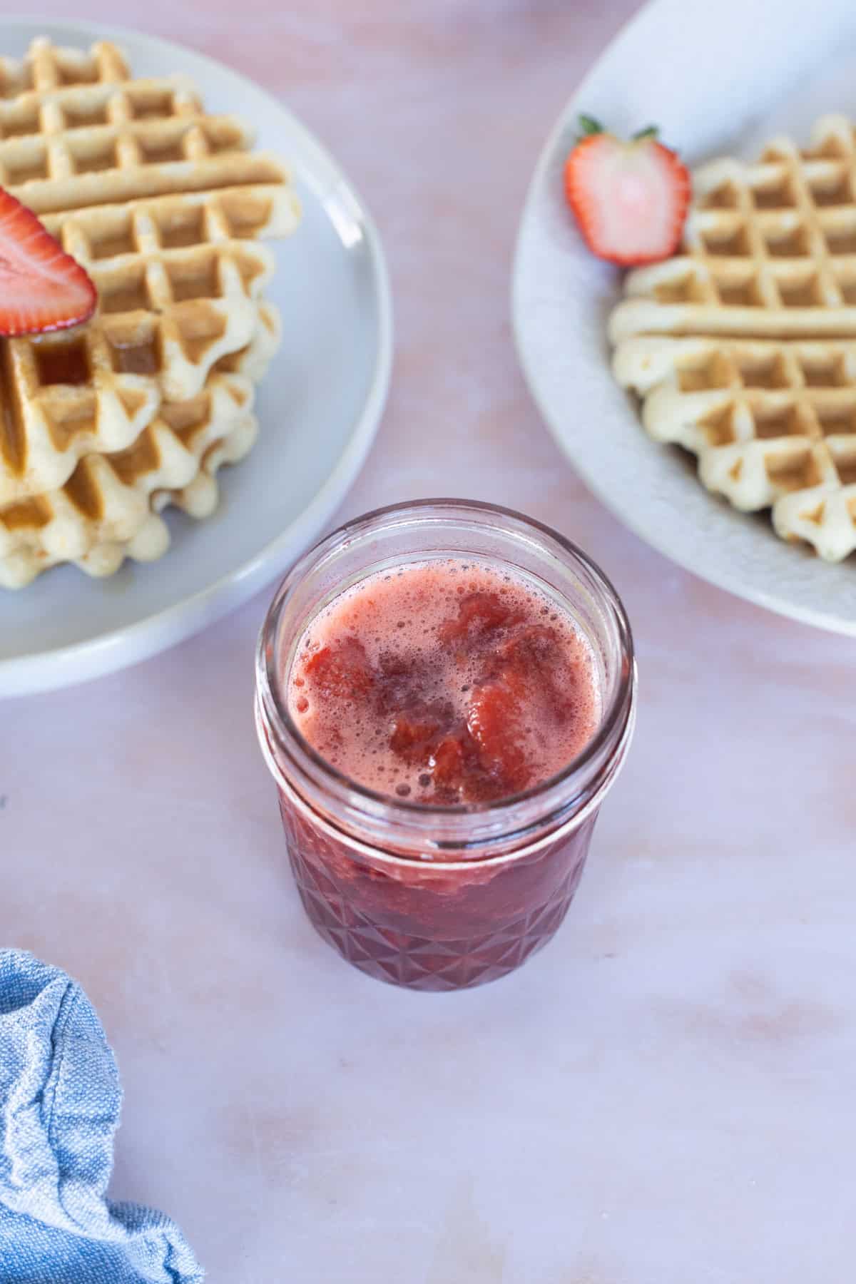 A glass jar of strawberry juice is placed on a pink surface, with two plates of waffles and sliced strawberries nearby.