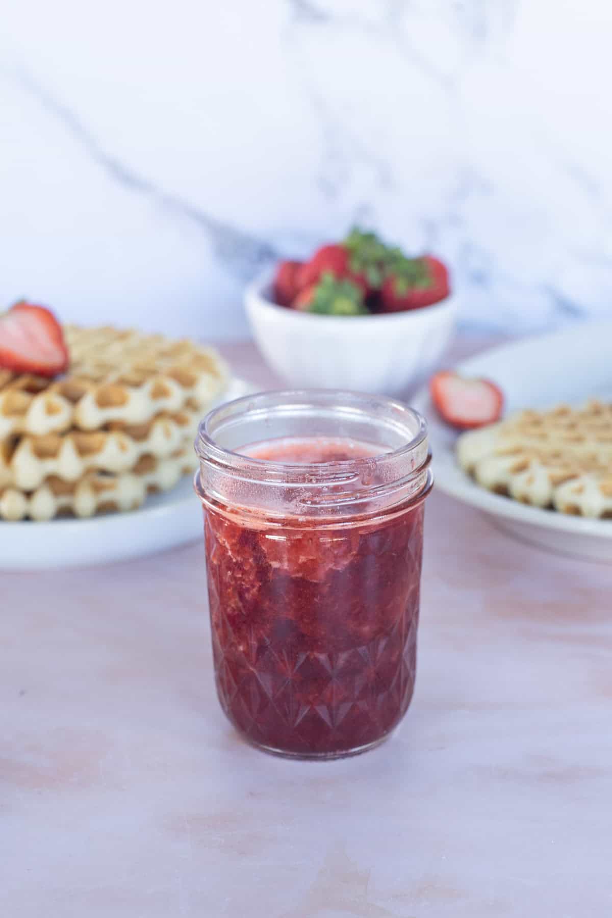 A glass jar of strawberry jam sits on a pink surface, with plates of waffles and a bowl of strawberries in the background.