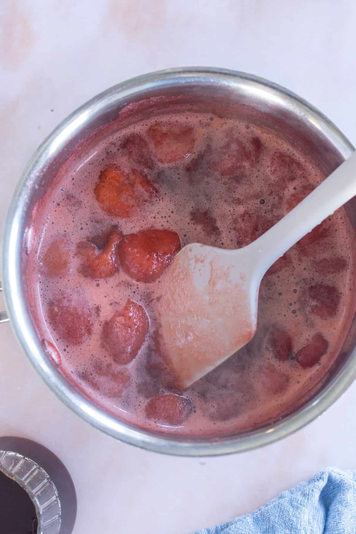 A pot of simmering strawberry mixture is being stirred with a white spatula. The mixture appears thick and chunky, suggesting it is being cooked down for jam or sauce.