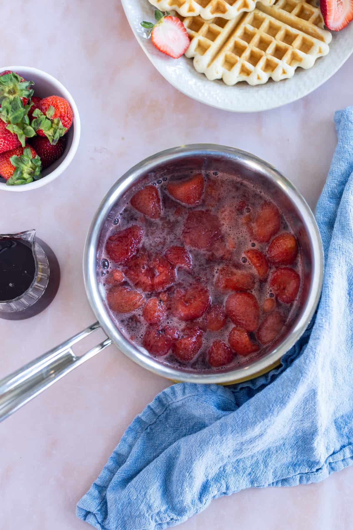 A saucepan filled with simmering strawberries sits on a counter near a blue cloth, with waffles and fresh strawberries visible in the background.