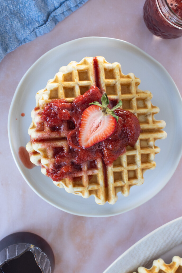 A plate with a waffle topped with strawberry sauce and a halved strawberry, shot from above on a light pink surface.
