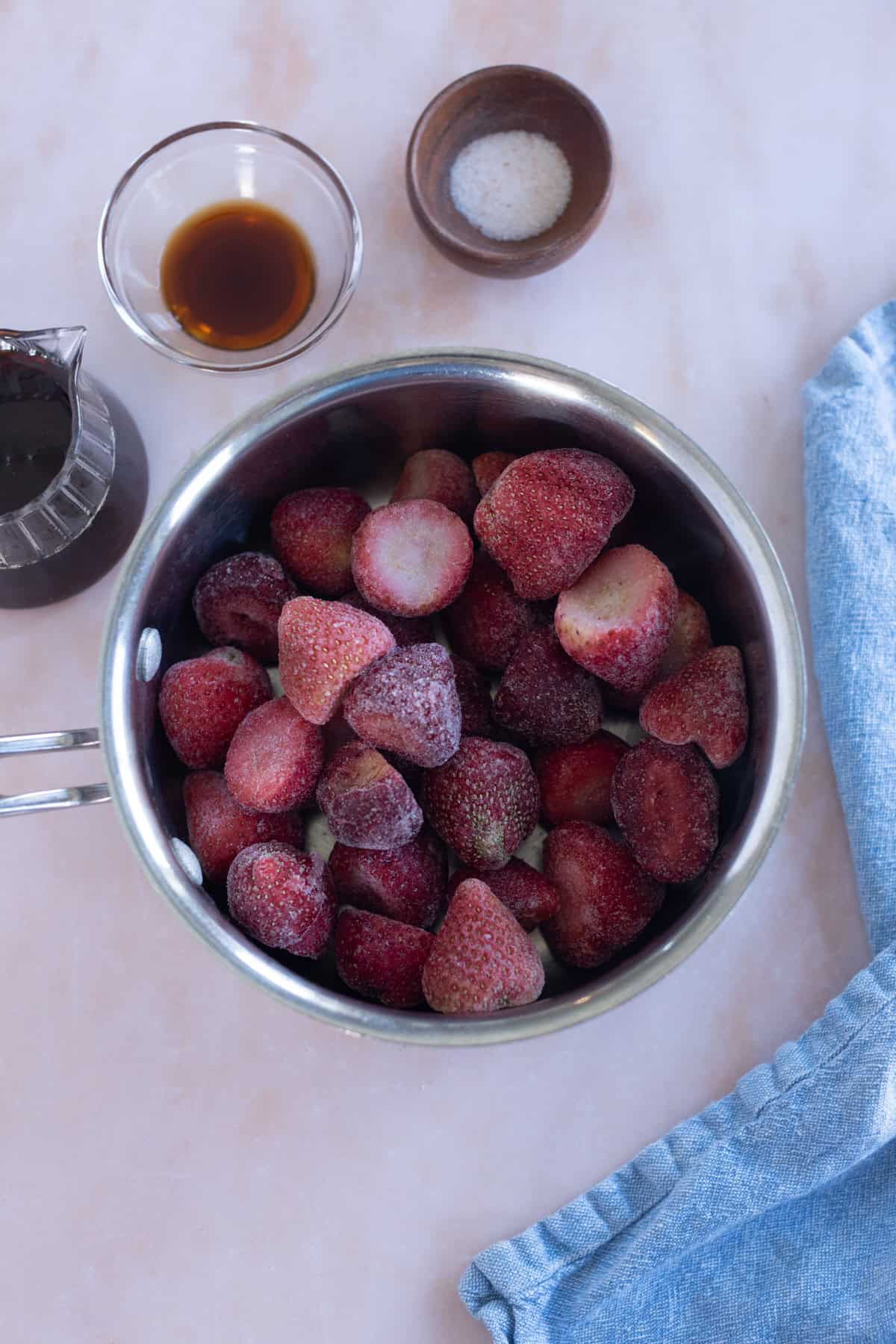 A pot filled with frozen strawberries sits on a counter next to a blue cloth, a bowl of salt, a bowl of vanilla extract, and a small pitcher of liquid.