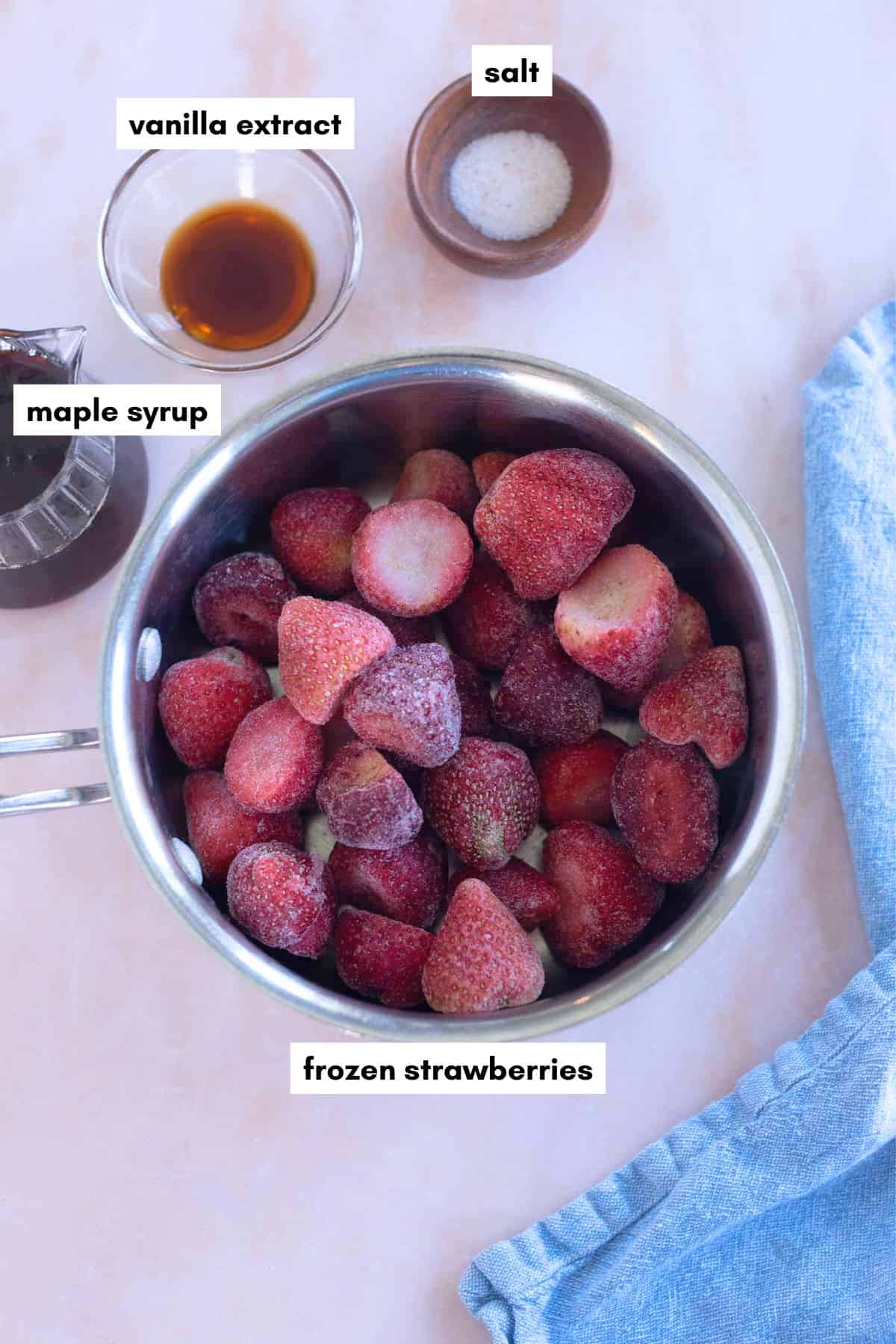 A saucepan of frozen strawberries destined for strawberry compote sits on a countertop, surrounded by bowls of maple syrup, vanilla extract, and salt, next to a blue cloth.