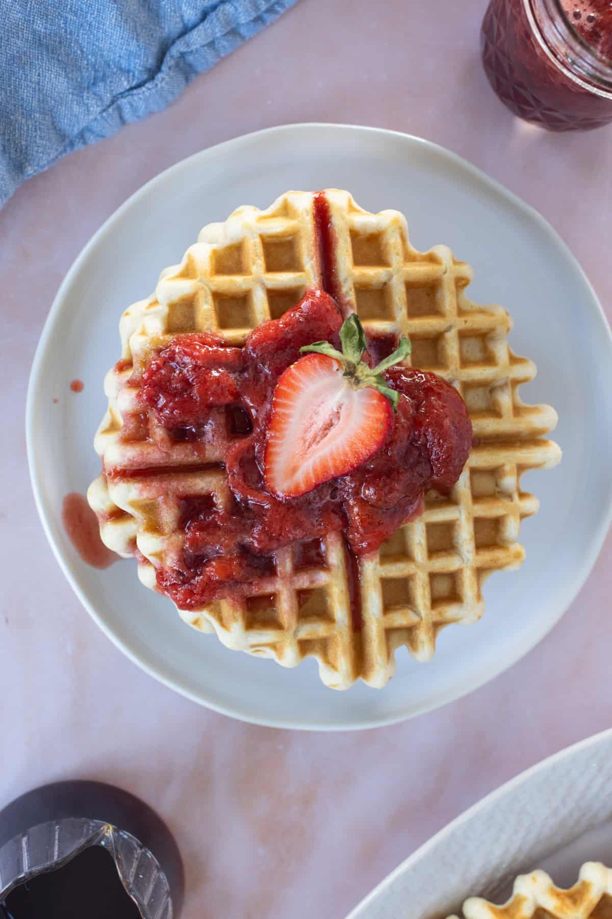 A plate with a waffle topped with strawberry sauce and a halved strawberry, shot from above on a light pink surface.