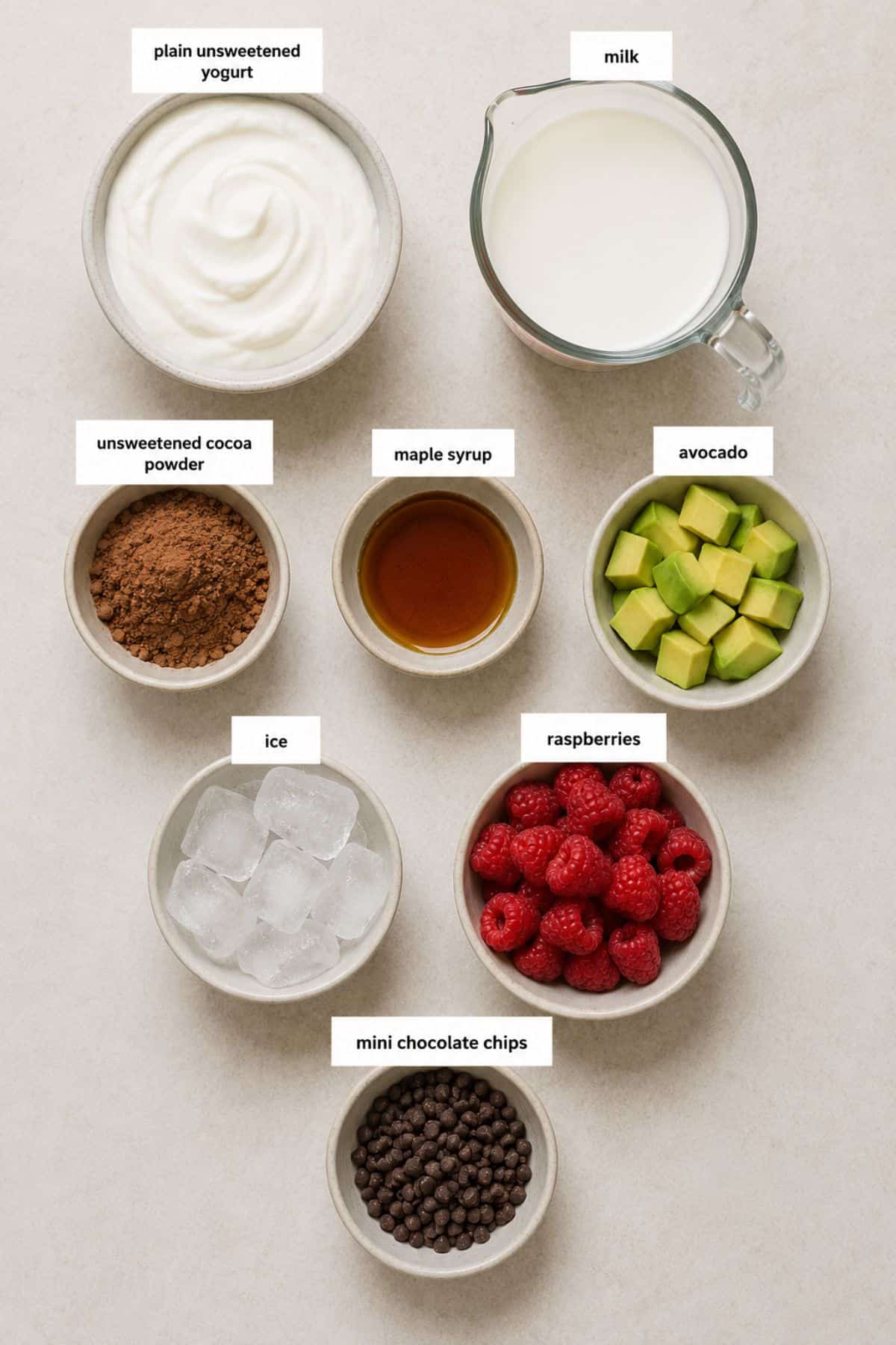 Bowls of yogurt, milk, cocoa powder, maple syrup, avocado, ice, raspberries, and mini chocolate chips arranged on a countertop—each ingredient labeled with its name—provide everything you need for a delicious chocolate smoothie. This easy smoothie recipe with avocado promises a creamy texture and rich flavor perfect for any time of day.